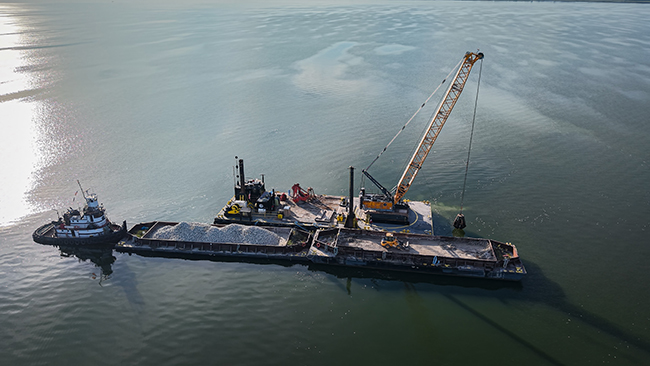 An aerial photo of a tug pushing two barges in a line that are loaded with cobble. Alongside the cobble barges is a third barge with a tall crane. The crane has a grapple on the end of the cable, hanging close to the water. The tug and barges are surrounded by calm water with the sun reflected on the left side.